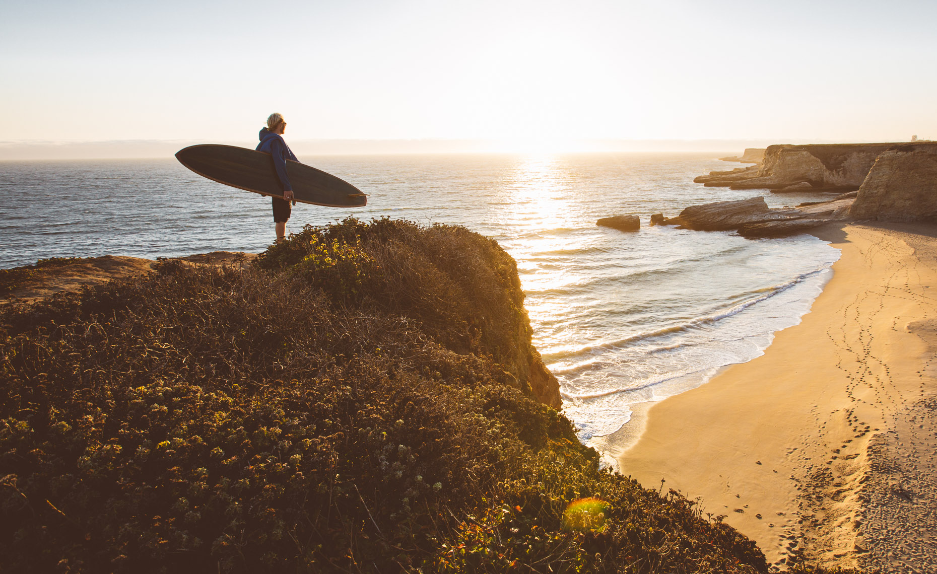 A surfer standing at the edge of a cliff overlooking the surf break at the beach, carrying a wooden longboard built by Martijn Stiphout from Ventana Surfboards