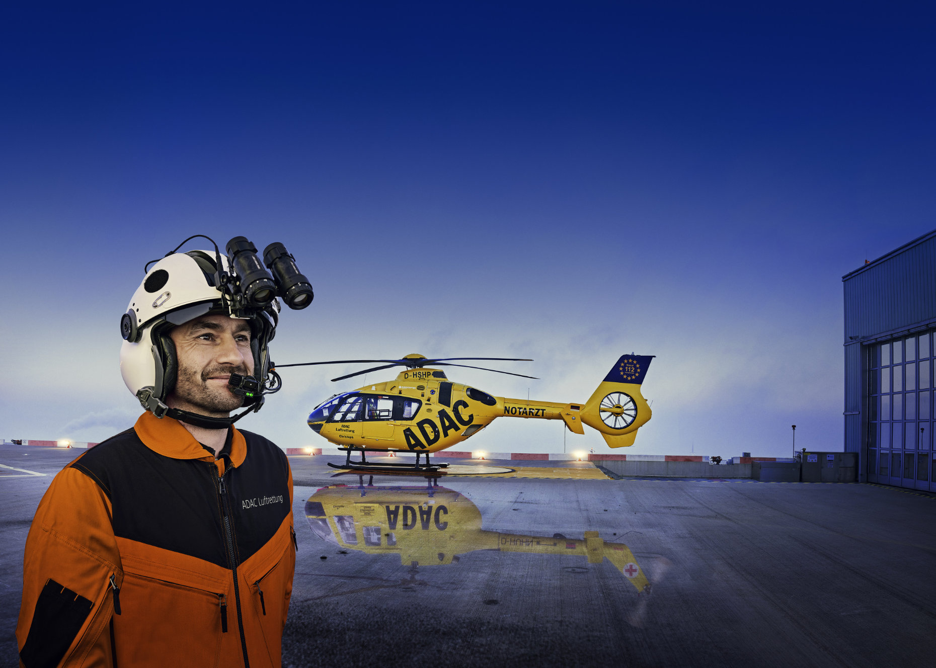Ein männlicher Helikopter Pilot mit Nachtsichtgeräte auf dem Helm steht vor einem Hubschrauber der ADAC Luftrettung auf dem Dach des Augsburger Krankenhaus