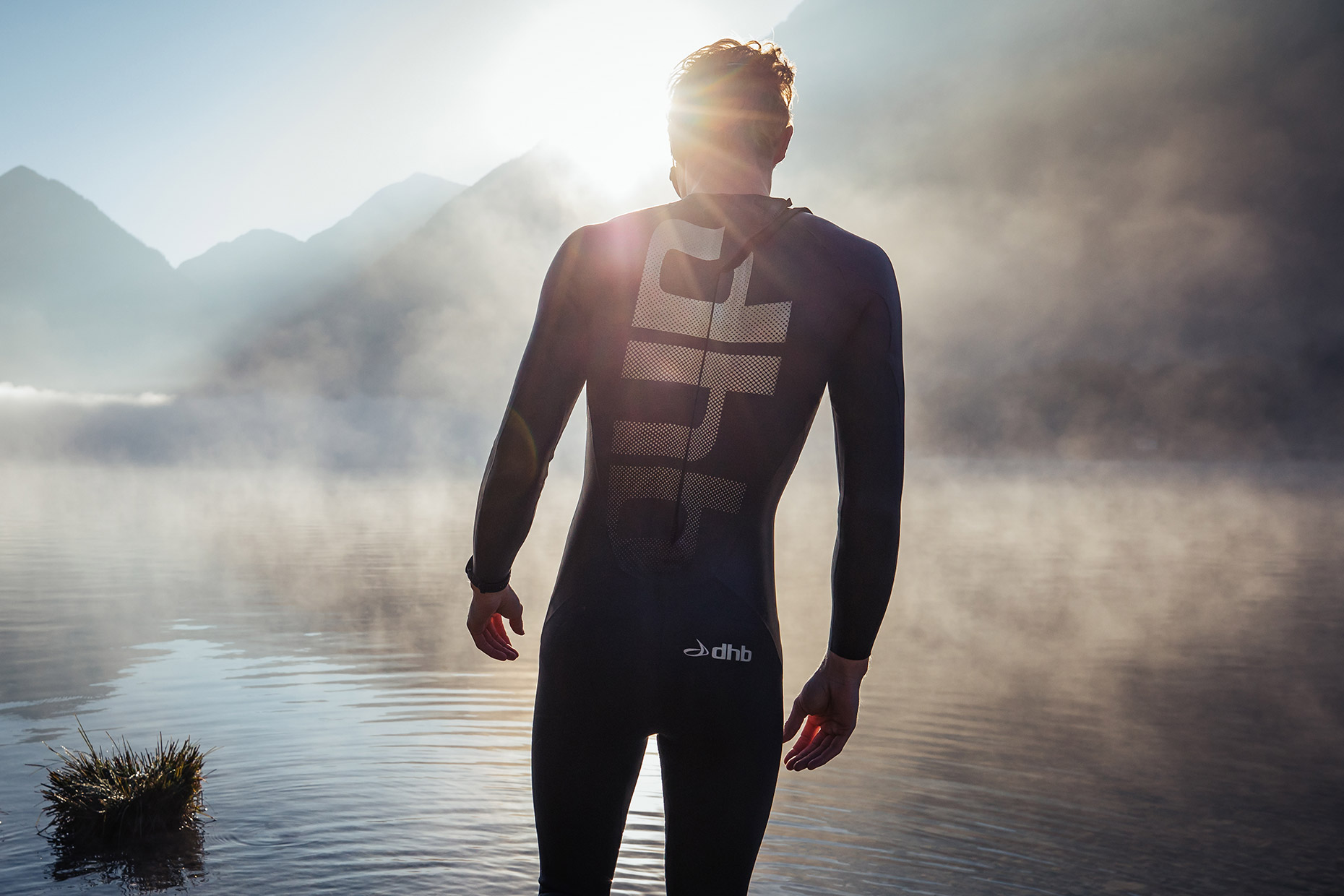 Male triathlete wearing a wetsuit standing on the edge of a lake at sunrise