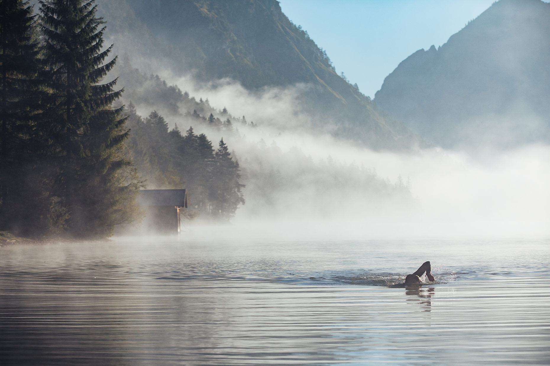 Early morning fog rising up from an alpine lake just after sunrise, a triathlete swimming in the distance 