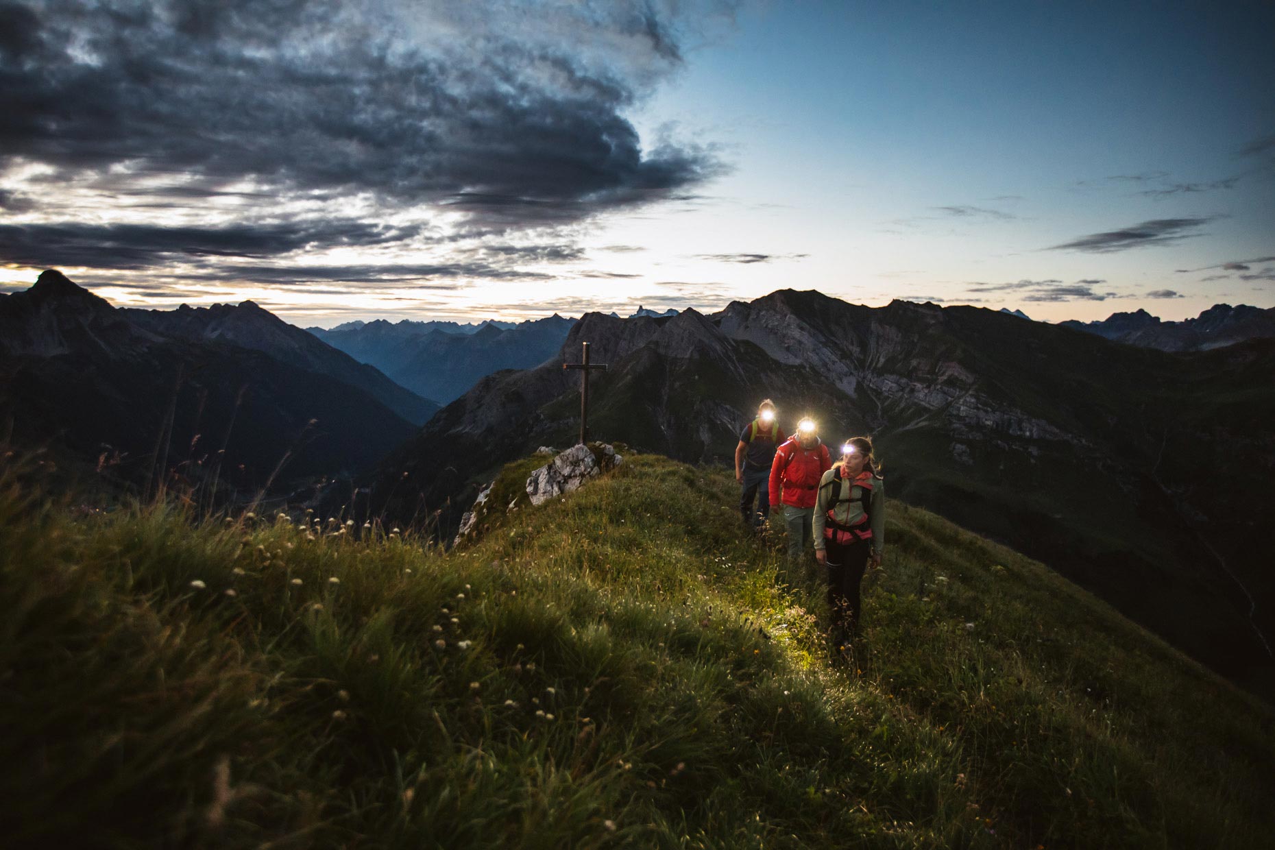Three hikers wearing headlamps hike along a mountain ridge in the dawn light just before sunrise
